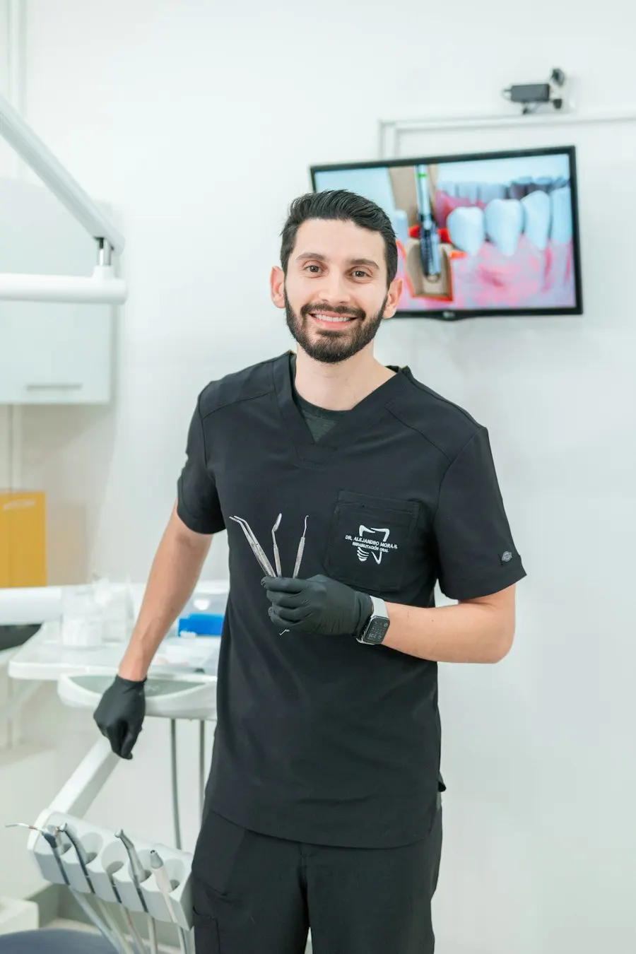 Dr. Daniel Aziz in dark scrubs holding dental instruments in a treatment room