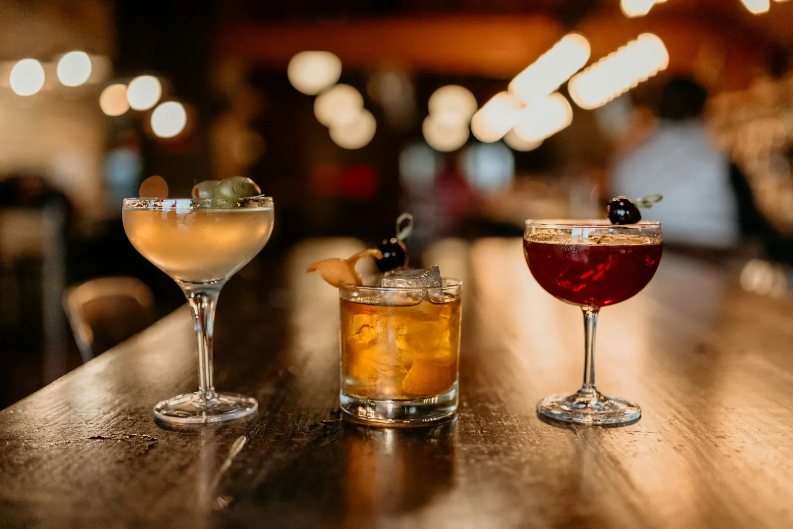 Three cocktails lined up on the bar with warm lights behind