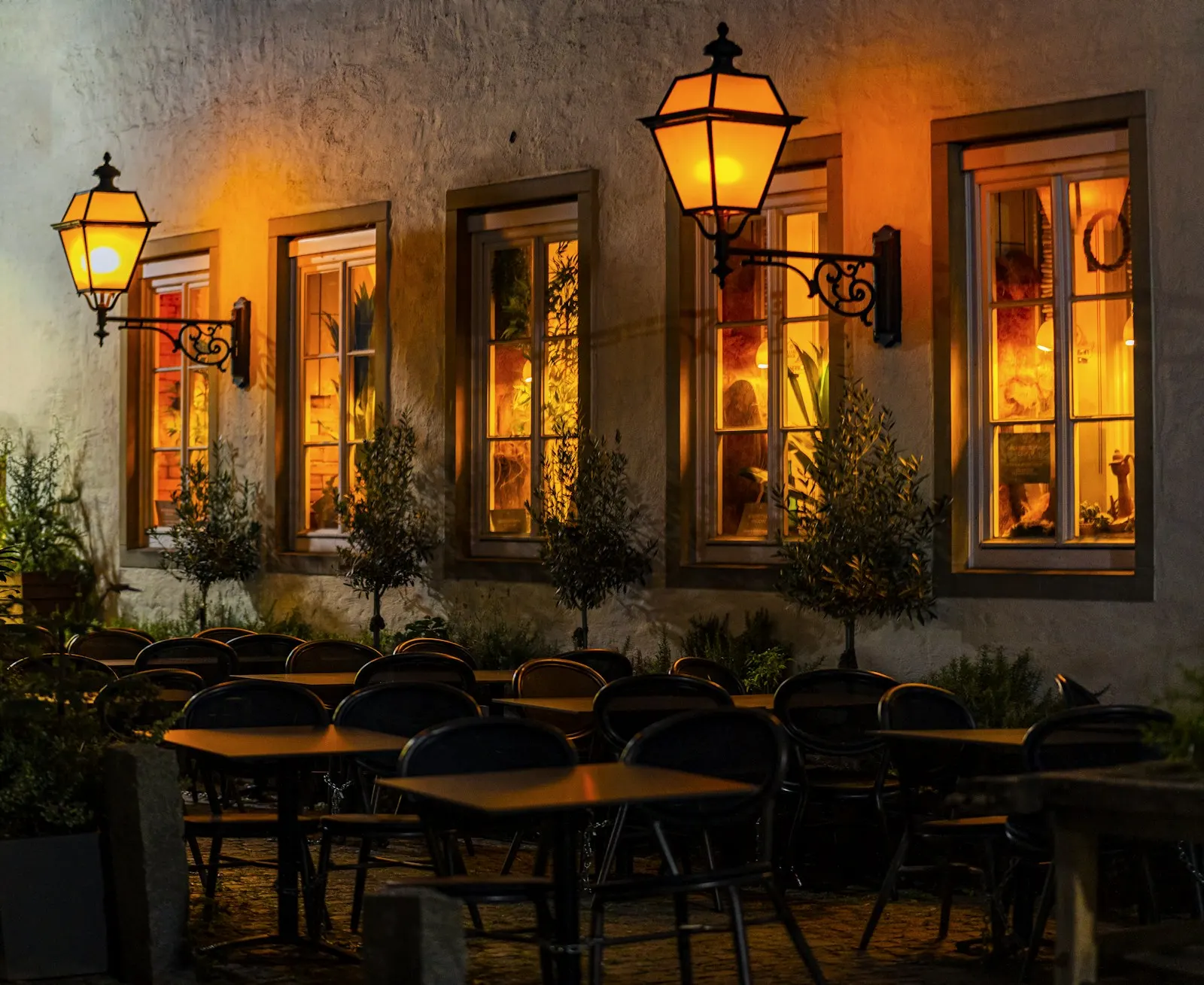 The restaurant’s front windows glowing under lanterns at night, with the patio set