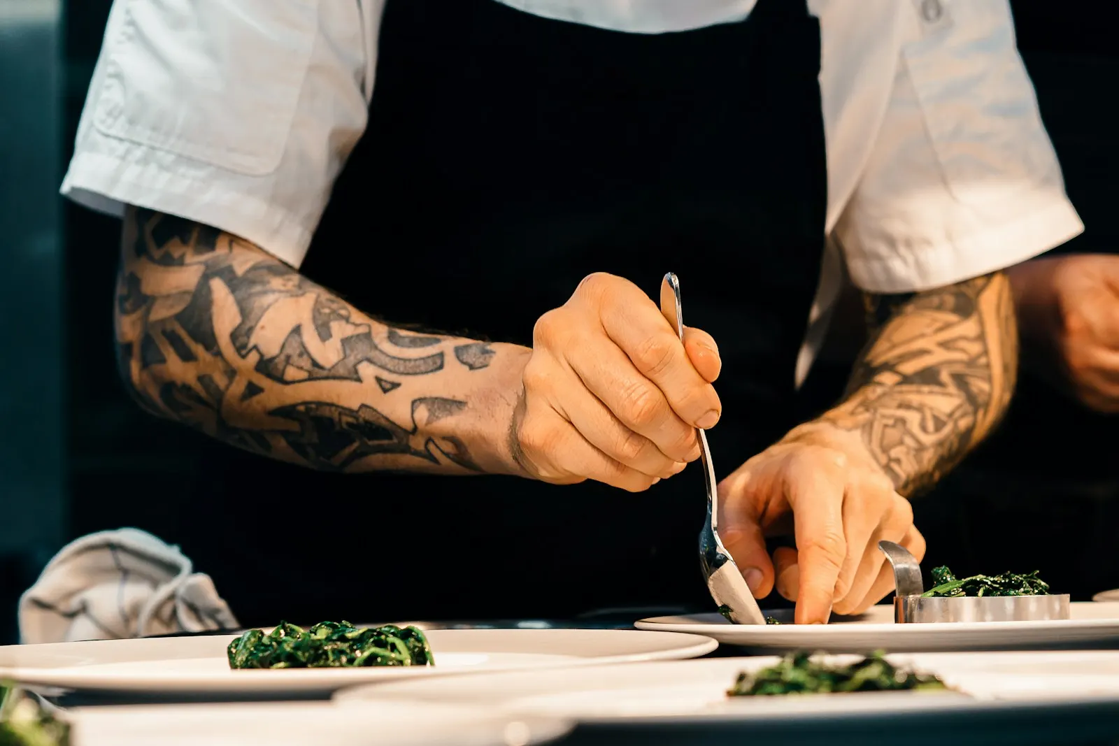 A cook with tattooed forearms plating greens at the pass