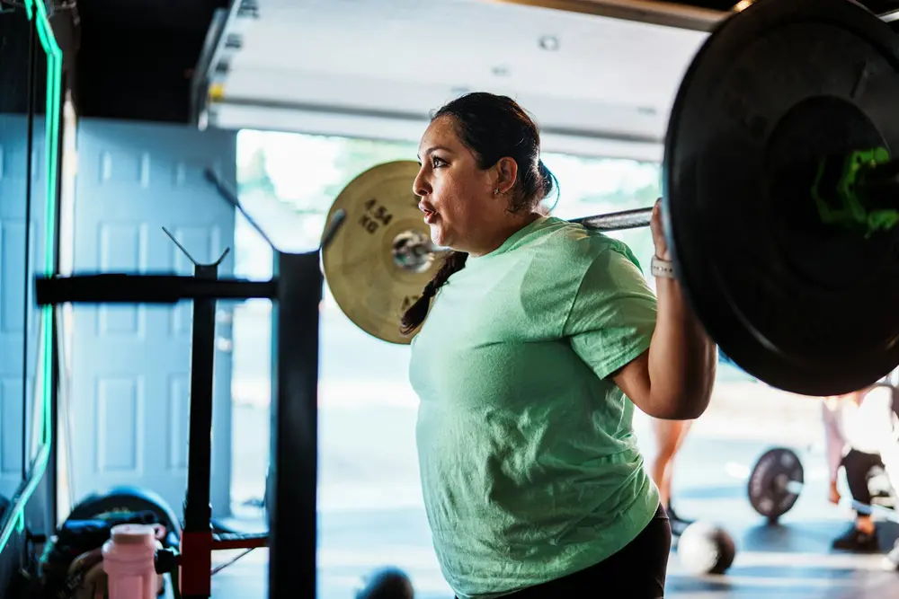 Denise, a member in a green T-shirt, back squatting a loaded barbell by the open garage door