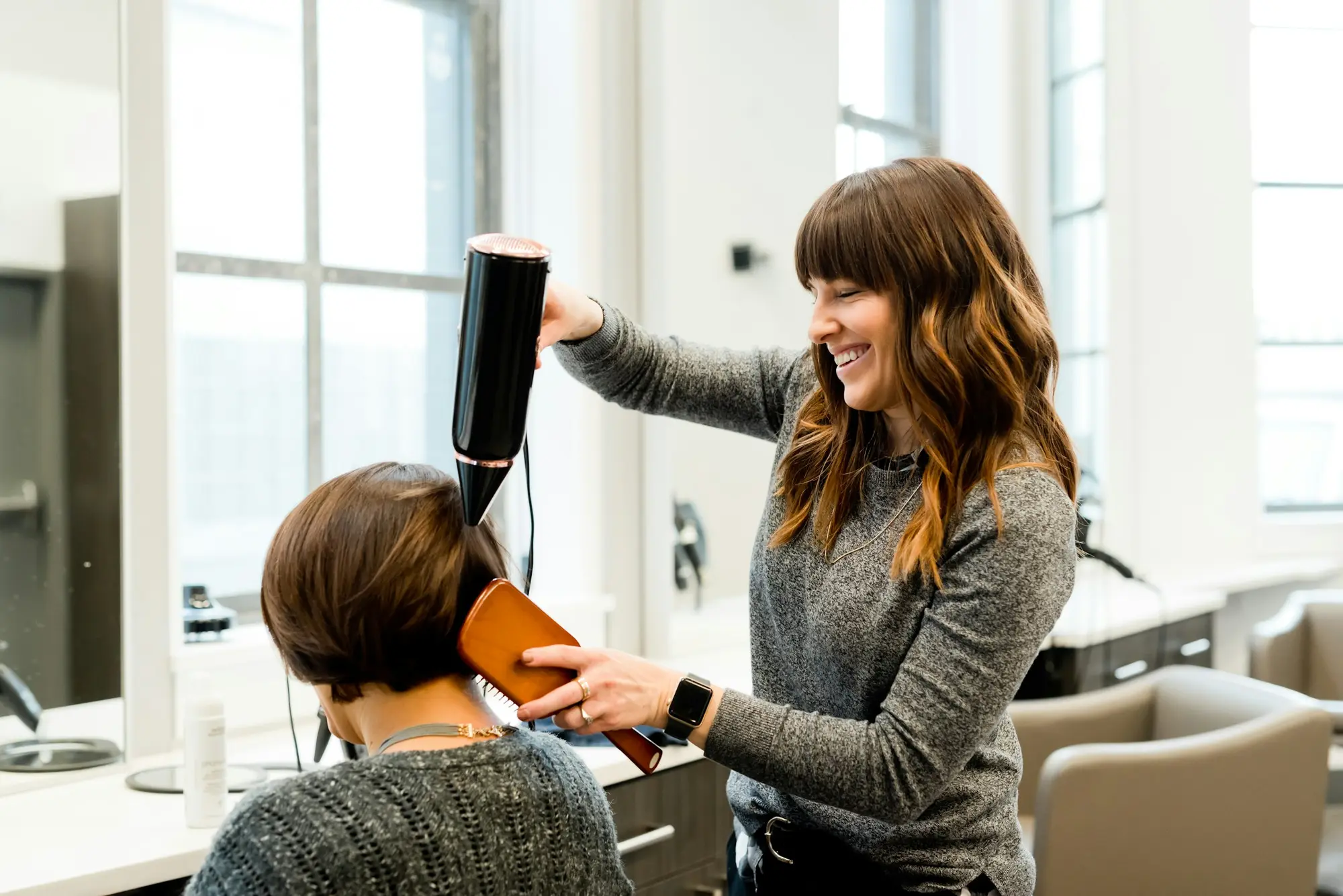 Hannah, smiling, blow-drying a client’s short brown hair in the bright salon
